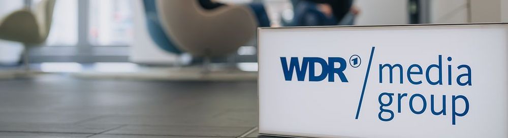 Ein Leuchtkasten mit dem WDR mediagroup Logo steht im Vordergrund. Hinten sitzen Menschen in Sesseln. | Bildquelle: WDR mediagroup GmbH Ein Leuchtkasten mit dem WDR mediagroup Logo steht im Vordergrund. Hinten sitzen Menschen in Sesseln.