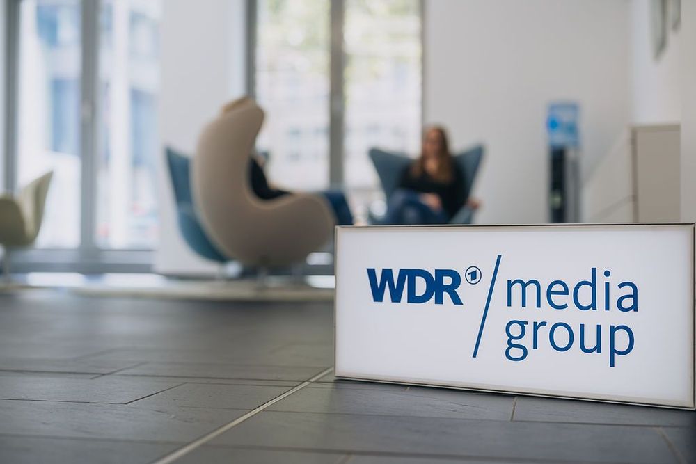 Ein Leuchtkasten mit dem WDR mediagroup Logo steht im Vordergrund. Hinten sitzen Menschen in Sesseln. | Bildquelle: WDR mediagroup GmbH Ein Leuchtkasten mit dem WDR mediagroup Logo steht im Vordergrund. Hinten sitzen Menschen in Sesseln.